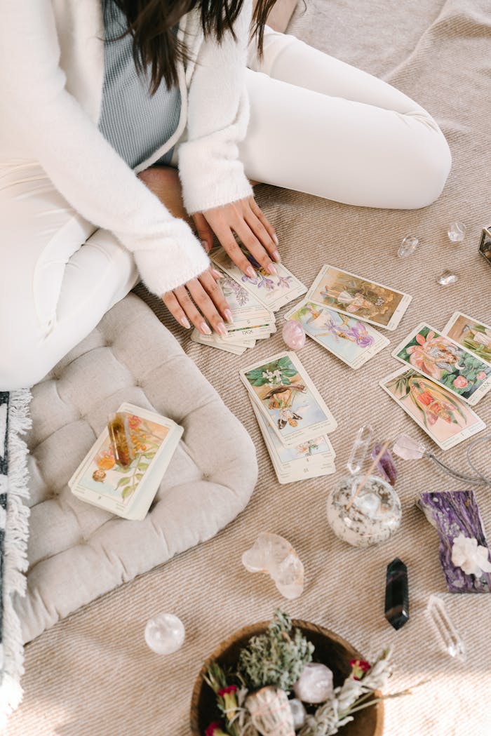 who-we-are A woman sits cross-legged indoors, handling tarot cards, surrounded by crystals and herbs.