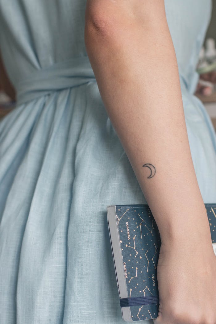 why-choose-us A woman's arm with a crescent moon tattoo holding a book in a blue dress.